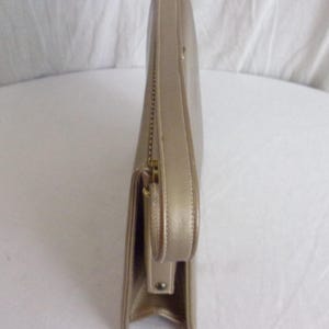 May include: A side view of a gold-colored clutch purse. The purse has a rectangular shape with a flap closure and a small handle. A gold zipper is visible along the side. The purse appears to be made of leather or a similar material.