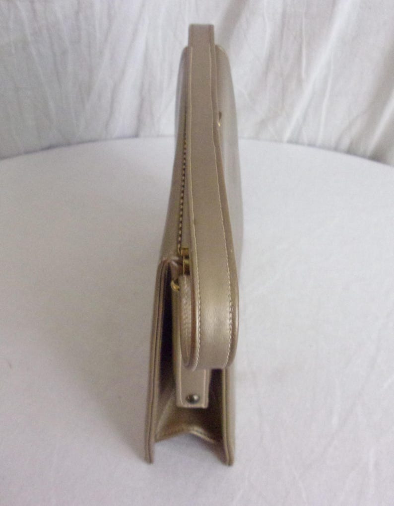 May include: A side view of a gold-colored clutch purse. The purse has a rectangular shape with a flap closure and a small handle. A gold zipper is visible along the side. The purse appears to be made of leather or a similar material.