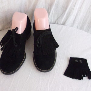 May include: Pair of black suede shoes with tassel detailing and a separate fringed accessory. The shoes have a rounded toe and a low heel. The accessory has two snap closures. The shoes are displayed on pink shoe forms.