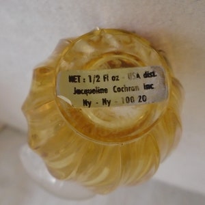 May include: A vintage, clear, amber-colored perfume bottle with a swirled design. The bottle has a label that reads "NET: 1/2 Fl oz - USA dist. Jacqueline Cochran Inc. Ny - Ny - 100 20."