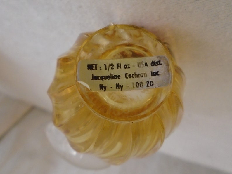 May include: A vintage, clear, amber-colored perfume bottle with a swirled design. The bottle has a label that reads "NET: 1/2 Fl oz - USA dist. Jacqueline Cochran Inc. Ny - Ny - 100 20."