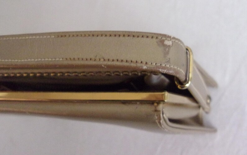 May include: A beige handbag with a gold-colored clasp and strap. The bag's strap is stitched and shows some wear. The clasp is rectangular and the bag appears to be made of a smooth material.