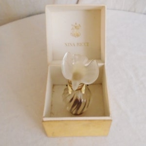 May include: A vintage Nina Ricci perfume bottle in its original box. The bottle has a clear, flower-shaped stopper and a swirled glass body. The box is cream-colored with the Nina Ricci logo printed on the inside.