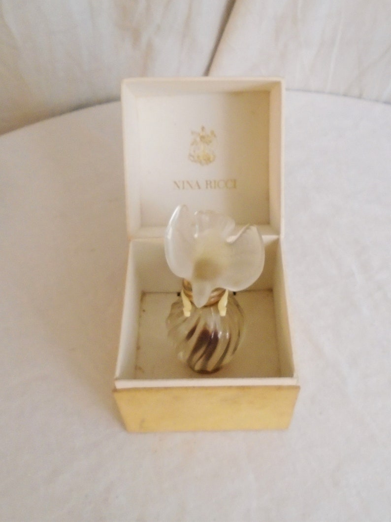 May include: A vintage Nina Ricci perfume bottle in its original box. The bottle has a clear, flower-shaped stopper and a swirled glass body. The box is cream-colored with the Nina Ricci logo printed on the inside.