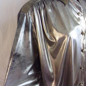 May include: Silver metallic button-down shirt with a subtle distressed finish. The long-sleeved shirt features a pointed collar and button closures. The fabric has a shiny, reflective quality, creating a vintage aesthetic.