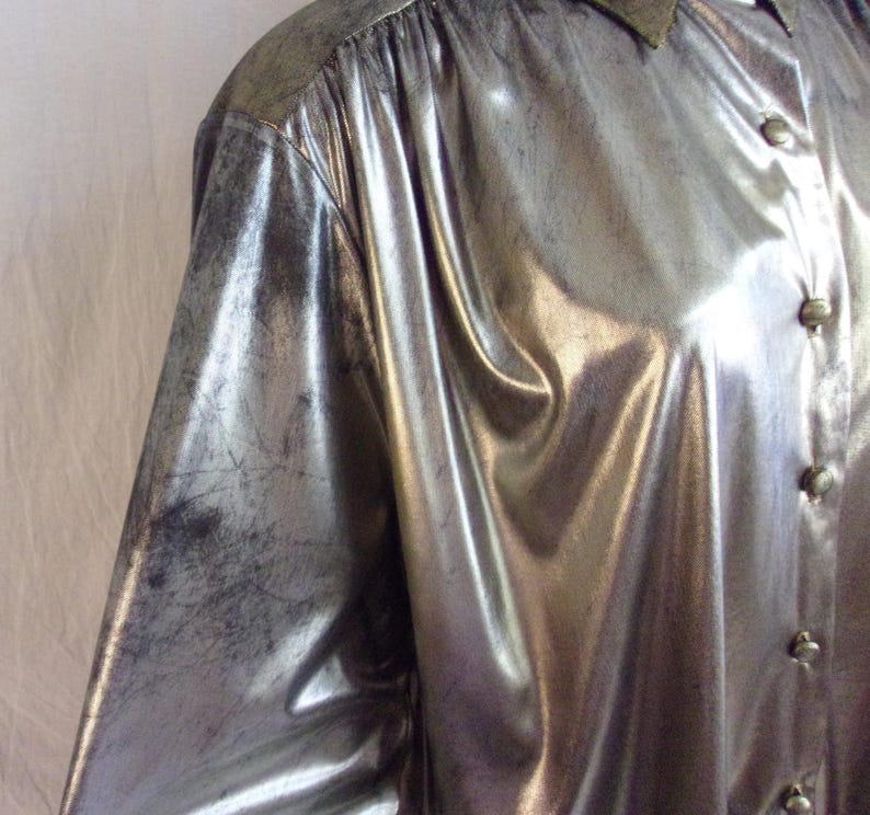 May include: Silver metallic button-down shirt with a subtle distressed finish. The long-sleeved shirt features a pointed collar and button closures. The fabric has a shiny, reflective quality, creating a vintage aesthetic.