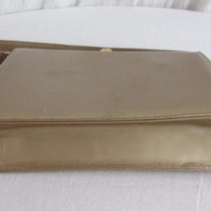 May include: A vintage gold-colored clutch purse with a matching strap. The rectangular bag has a simple, elegant design with a fold-over flap and a subtle sheen. The strap is thin and attached to the top of the purse.