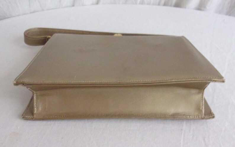 May include: A vintage gold-colored clutch purse with a matching strap. The rectangular bag has a simple, elegant design with a fold-over flap and a subtle sheen. The strap is thin and attached to the top of the purse.