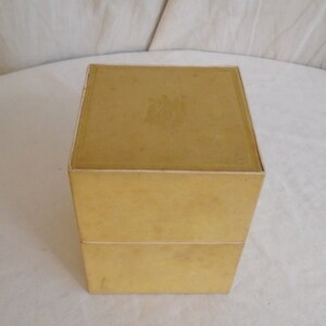May include: A vintage, two-piece, square box in a muted yellow color. The box has a simple design with a slightly textured surface. The top of the box has a faded emblem. The box is sitting on a white surface.