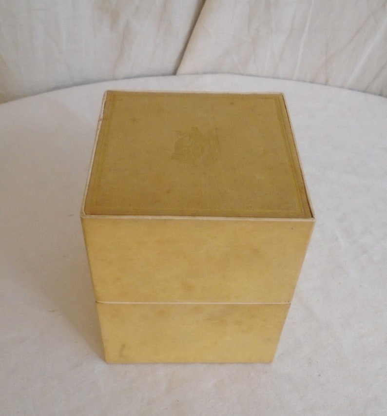 May include: A vintage, two-piece, square box in a muted yellow color. The box has a simple design with a slightly textured surface. The top of the box has a faded emblem. The box is sitting on a white surface.