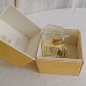 May include: A vintage perfume bottle with a frosted flower-shaped stopper, presented in a cream-colored box. The bottle is clear with a ribbed design. The box has a gold exterior and a cream interior with the brand name.