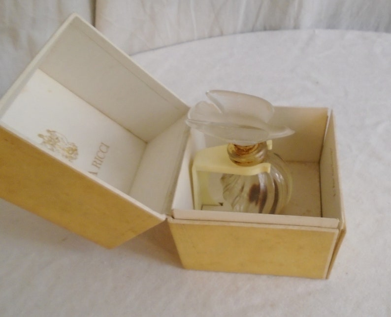 May include: A vintage perfume bottle with a frosted flower-shaped stopper, presented in a cream-colored box. The bottle is clear with a ribbed design. The box has a gold exterior and a cream interior with the brand name.