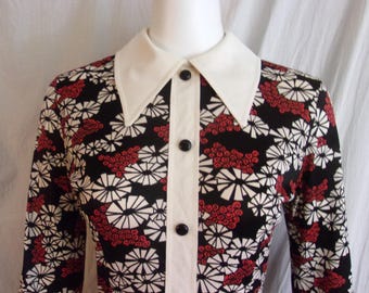 Puede incluir: Una camisa de manga larga con fondo negro y un patrón repetitivo de formas florales geométricas blancas y motivos rojos en forma de nubes. La camisa tiene un cuello puntiagudo blanco sólido y una tapeta central blanca con botones negros.