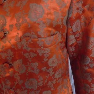May include: A close-up of a bright orange jacket with a floral pattern. The jacket has a mandarin collar and a button closure. The fabric is shiny and has a subtle sheen.