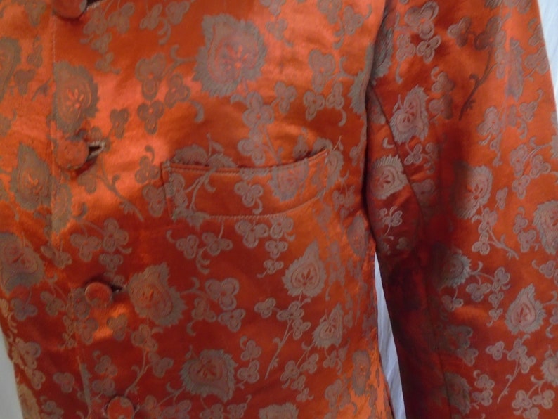 May include: A close-up of a bright orange jacket with a floral pattern. The jacket has a mandarin collar and a button closure. The fabric is shiny and has a subtle sheen.