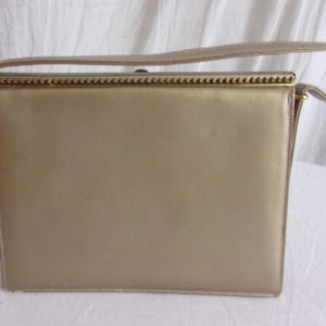 May include: A rectangular, gold-colored handbag with a matching handle. The bag has a structured design with a clasp closure and gold-toned metal accents. The handle is a flat, fabric strap.