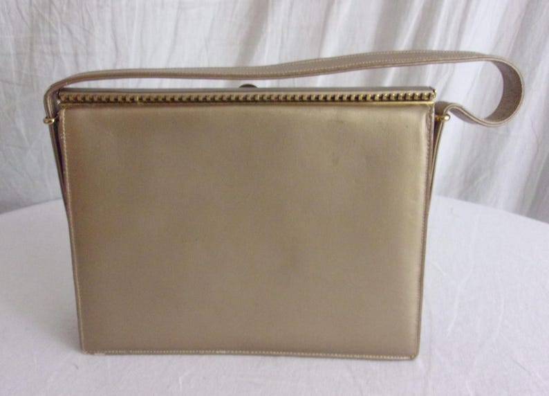 May include: A rectangular, gold-colored handbag with a matching handle. The bag has a structured design with a clasp closure and gold-toned metal accents. The handle is a flat, fabric strap.