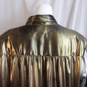 May include: A metallic gold shirt with a shiny, textured surface. The shirt has a collar and a pleated detail at the back. The fabric has a distressed, vintage appearance.
