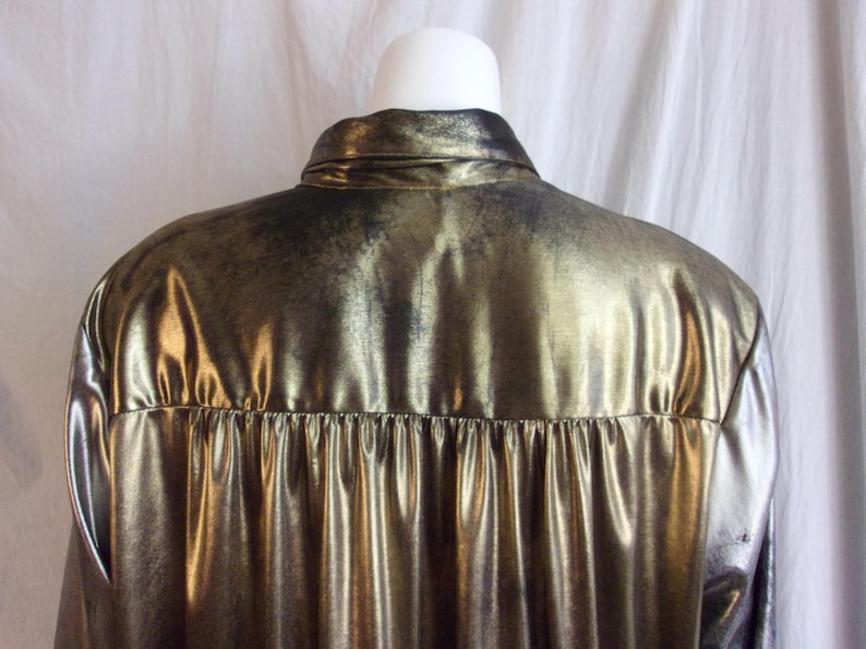 May include: A metallic gold shirt with a shiny, textured surface. The shirt has a collar and a pleated detail at the back. The fabric has a distressed, vintage appearance.