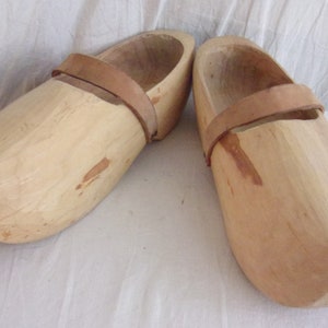 Könnte beinhalten: Ein Paar Holzschuhe mit Lederriemen. Die Clogs sind hellbraun und haben ein schlichtes Design.