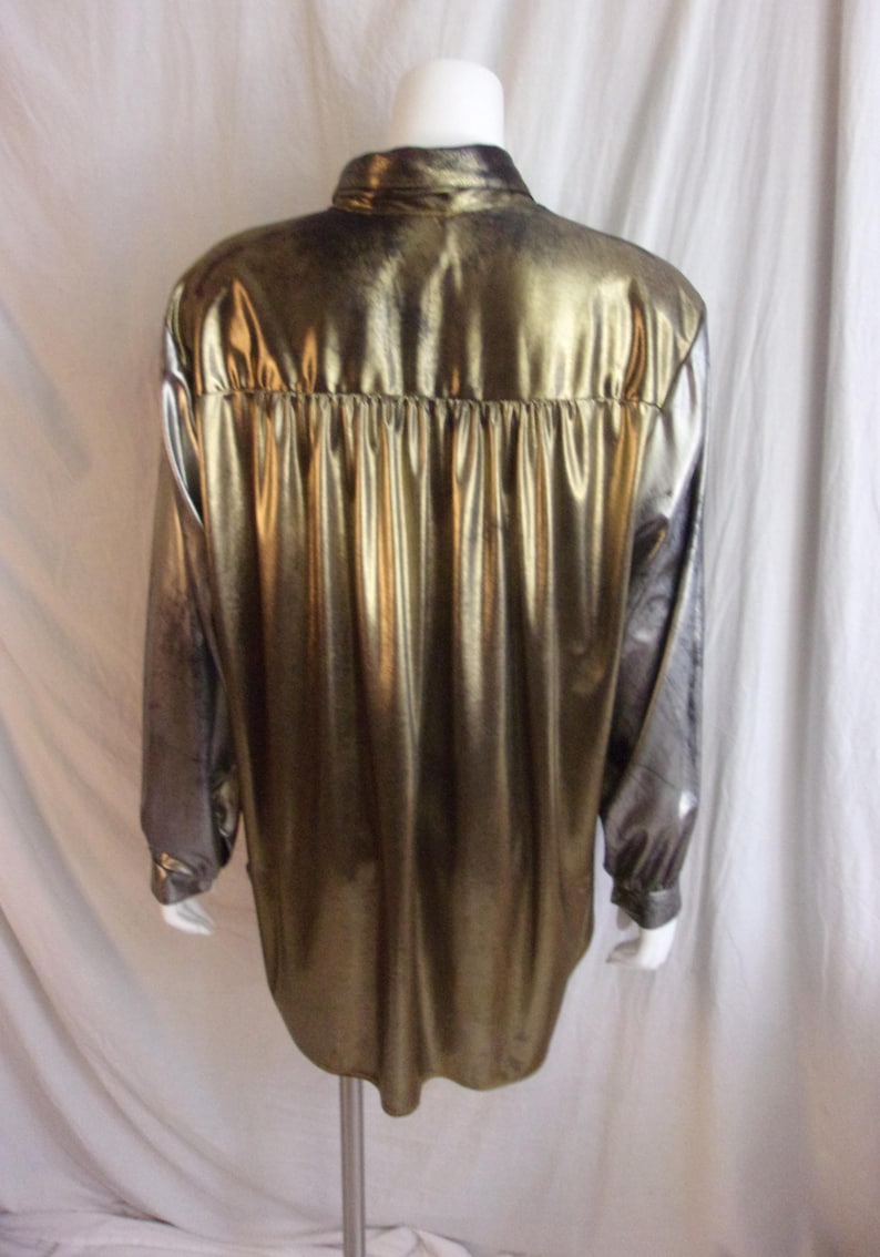 May include: A metallic gold long-sleeve shirt with a button-down collar. The shirt has a shiny, reflective surface and a gathered detail at the back yoke. The sleeves transition to a darker, greyish tone.