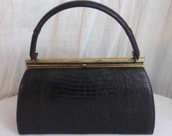 May include: A black handbag with a textured lizard-skin pattern, featuring a single arched handle and a rectangular gold-tone metal frame closure. The bag has a structured, semi-circular shape with a flat base and a hinged top opening mechanism.
