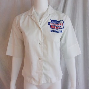 May include: White short-sleeved button-up shirt with a blue and red patch that reads "National Cheerleaders Association Instructor".