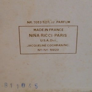 May include: A vintage perfume box with text that reads "NR. 7053 1/2 fl. oz. PARFUM" and "MADE IN FRANCE NINA RICCI PARIS." The box is off-white with a rectangular label in a darker shade. The bottom has the number "B11045."
