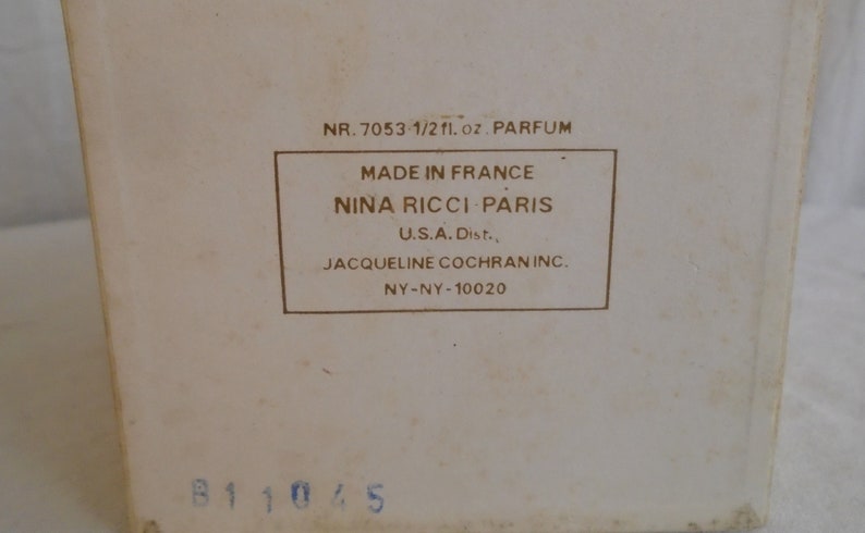 May include: A vintage perfume box with text that reads "NR. 7053 1/2 fl. oz. PARFUM" and "MADE IN FRANCE NINA RICCI PARIS." The box is off-white with a rectangular label in a darker shade. The bottom has the number "B11045."