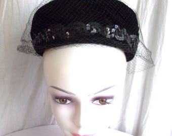 May include: Black velvet pillbox hat with a delicate black netting veil. The hat is adorned with sequins and small gold-colored beads. A vintage-inspired accessory.