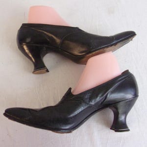 Peut inclure: Une paire de chaussures en cuir noir avec des bouts pointus et des talons incurv&eacute;s. Les chaussures sont pr&eacute;sent&eacute;es sur des formes &agrave; chaussures roses, mettant en valeur leur style vintage. Les talons mesurent environ 5 cm de haut.