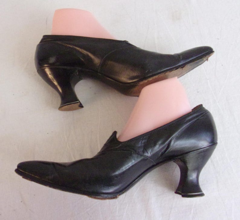Peut inclure: Une paire de chaussures en cuir noir avec des bouts pointus et des talons incurv&eacute;s. Les chaussures sont pr&eacute;sent&eacute;es sur des formes &agrave; chaussures roses, mettant en valeur leur style vintage. Les talons mesurent environ 5 cm de haut.