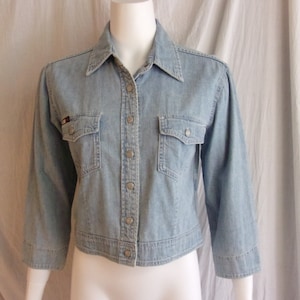 May include: A light blue denim shirt with a button-up front, long sleeves, and two chest pockets. The shirt has a cropped length and a classic collar.