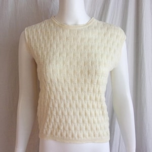 May include: Cream-colored sleeveless sweater with a textured, diamond-patterned design. The top has a crew neck and a slightly flared hem. The sweater is displayed on a mannequin against a white backdrop, showcasing its vintage style and knit construction.