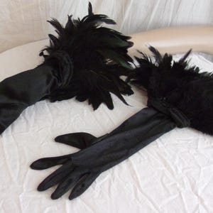 May include: A pair of black satin opera-length gloves featuring dense black feather trim at the cuffs. The gloves have a smooth, semi-matte finish and extend past the wrist, with the feathers creating a voluminous, textured accent at the opening.