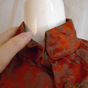 May include: A close-up of a red and gold brocade fabric with a floral pattern. The fabric is folded over to show the inside of the collar.
