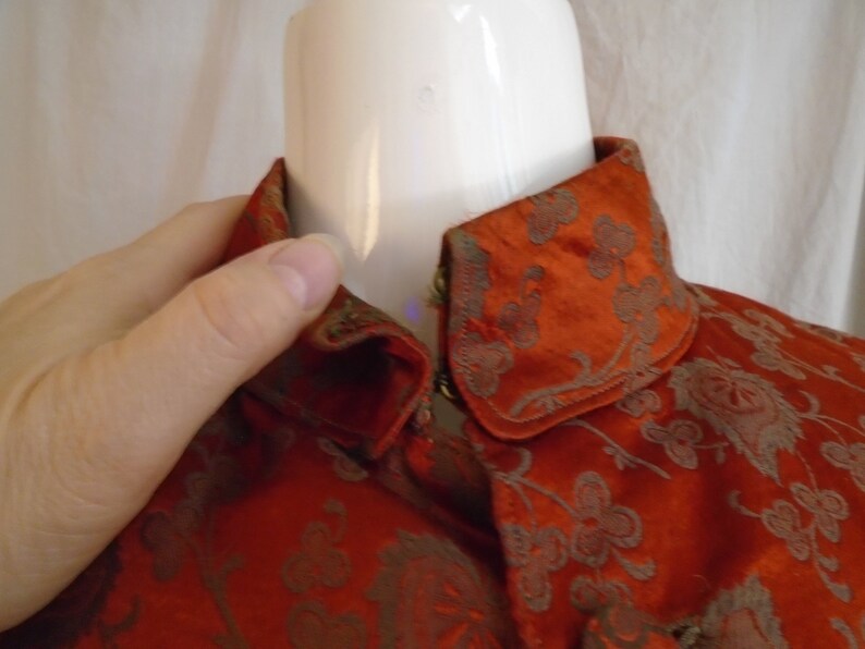 May include: A close-up of a red and gold brocade fabric with a floral pattern. The fabric is folded over to show the inside of the collar.