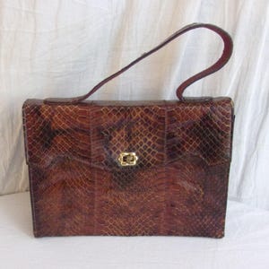 May include: A vintage brown handbag with a snakeskin pattern. The rectangular bag features a top handle and a gold-colored clasp. The bag's exterior has a textured appearance, with a flap closure and a single handle.