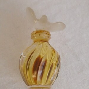 May include: A vintage perfume bottle with a swirling yellow glass body and a frosted white butterfly-shaped stopper. The bottle has a small, rounded shape and a cork closure. The glass has a subtle, swirled pattern.