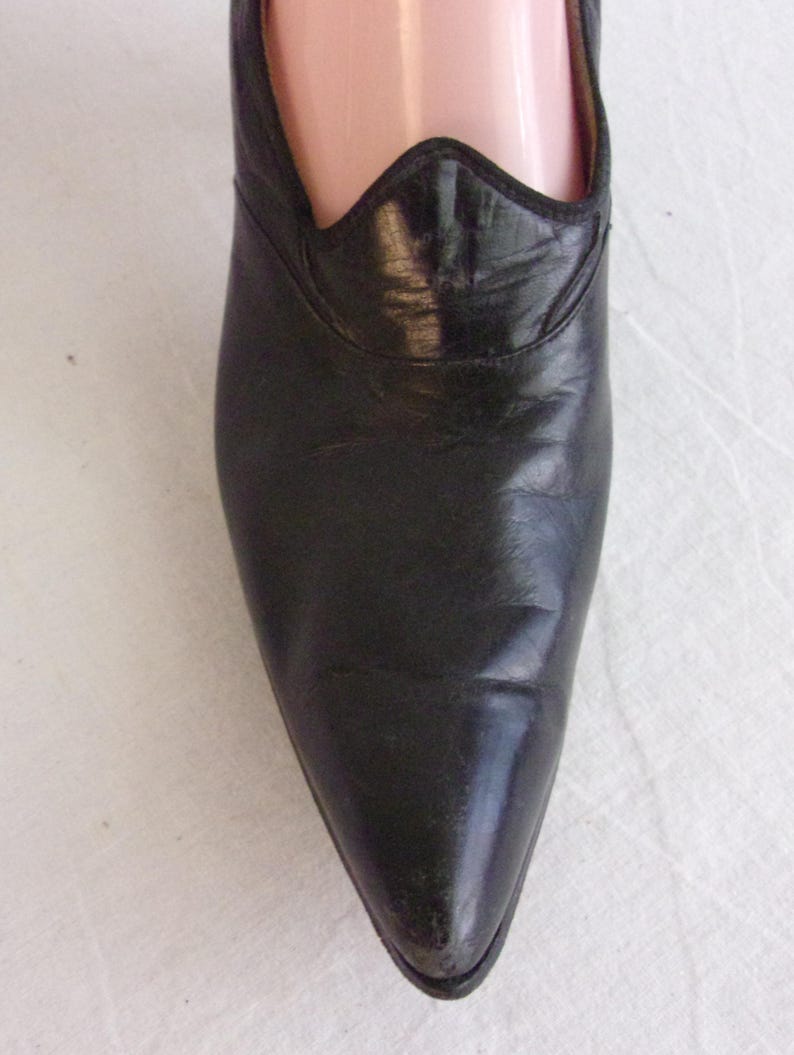 Peut inclure: Gros plan d'une chaussure en cuir noir &agrave; bout pointu. La chaussure pr&eacute;sente un bord d&eacute;coratif festonn&eacute; autour de l'ouverture. Le cuir semble l&eacute;g&egrave;rement us&eacute;, avec des plis visibles.