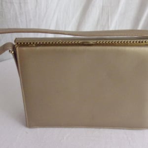 May include: A rectangular, gold-colored handbag with a curved handle. The bag has a structured design with a clasp closure and a stitched border. The handle is attached to the top of the bag.
