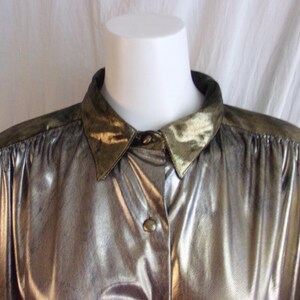 May include: A metallic gold button-down shirt with a pointed collar and shoulder accents. The shirt has long sleeves and a shiny, textured appearance. The collar and shoulder areas have a darker, olive-green hue.