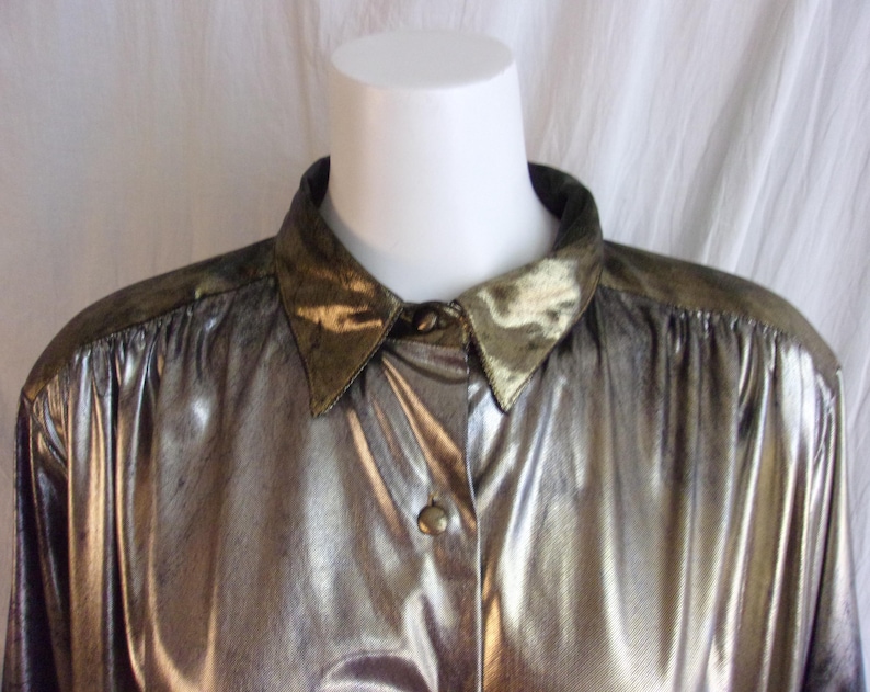 May include: A metallic gold button-down shirt with a pointed collar and shoulder accents. The shirt has long sleeves and a shiny, textured appearance. The collar and shoulder areas have a darker, olive-green hue.