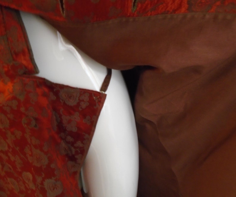 May include: A close-up of a red and gold brocade fabric with a brown lining. The fabric is draped over a white surface.