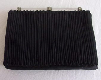 May include: A black pleated clutch purse with a rectangular shape. The purse has a silver-colored clasp with decorative rhinestones. The fabric is vertically pleated, creating a textured surface. The purse is likely vintage.