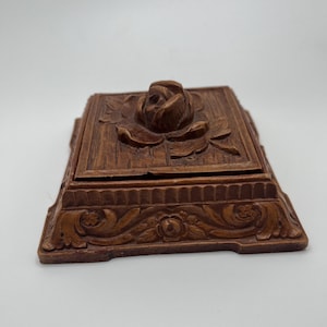 Puede incluir: Una caja de madera rectangular marrón con una rosa tallada en la tapa. La caja tiene un diseño floral detallado en la base y la tapa, con un borde decorativo ligeramente elevado. La caja es probablemente vintage o antigua.