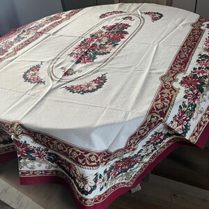 Puede incluir: Mantel crema con un diseño de flor de Pascua. El mantel presenta un óvalo floral central y bordes con detalles en rojo, verde y dorado. Las esquinas tienen arreglos florales adicionales, creando un tema navideño.