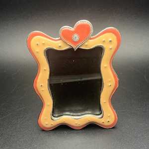 May include: A decorative picture frame with a heart-shaped accent. The frame has a wavy edge with a cream-colored interior, a red border, and small silver dots. A red heart with a silver border and a small gem sits at the top.
