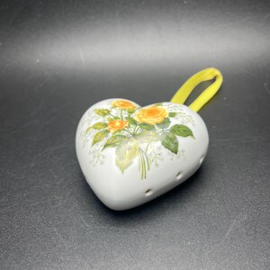 May include: White heart-shaped ceramic trinket box with a yellow ribbon handle. The box is decorated with a floral design featuring yellow roses and green leaves. The trinket box has several small holes on the side.