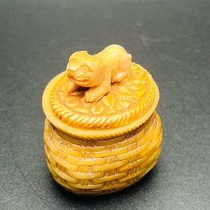 May include: A small, intricately carved basket with a lid, made of a light tan material. A small animal figure rests on the lid. The basket has a woven texture, and the lid features a detailed floral design. The overall aesthetic is rustic and artisanal.